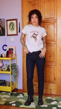 Oseba s kodrastimi lasmi, oblečena v belo polo majico Ralph Lauren, temne kavbojke in črne škornje, stoji v zaprtih prostorih na zeleni cvetlični preprogi ob lesenih vratih.