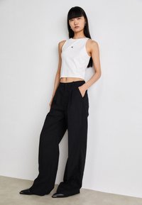 Bijela bezrukavna crop majica s crnim logotipom, uparena s crnim širokim hlačama visokog struka. Model stoji na neutralnoj površini.