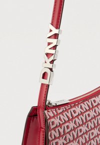 DKNY-laukku, jossa on punainen kiiltävän nahkan hihna, teksturoitu runko ja toistuva logokuvio, sekä hopeanvärisiä metalliosia.