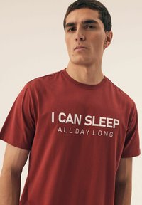 Czerwona bawełniana koszulka z krótkim rękawem i okrągłym dekoltem, z wyraźnym białym napisem: "MOGĘ SPAĆ CAŁY DZIEŃ." Gładka tekstura.