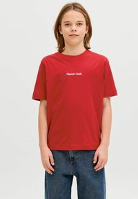 Vállig érő világosbarna hajú gyermek piros, fehér feliratos pólót és bő kék farmert visel, semleges, világos háttér előtt áll.