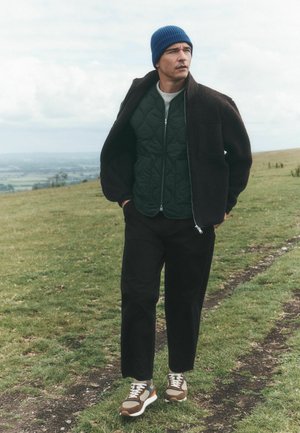 Man wearing a blue beanie, black fleece jacket, green quilted vest, black pants, and sneakers walking on a grassy hill with a cloudy sky.