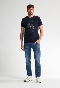 T-shirt in cotone blu navy con design grafico di una motocicletta. Abbinato a jeans di denim blu e sneakers bianche. Scollo rotondo semplice.