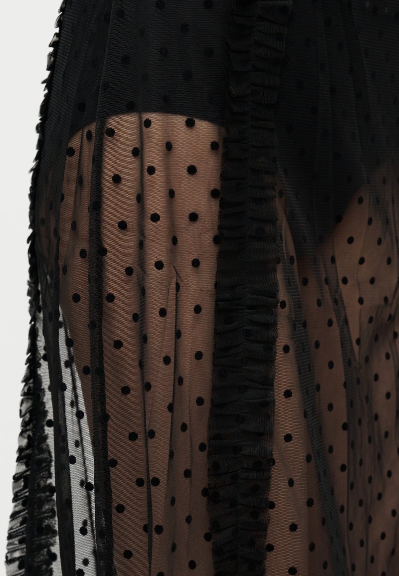 Tissu transparent à pois noirs superposé sur une base couleur nude, avec des bords volantés. La texture est douce avec une apparence délicate et transparente.