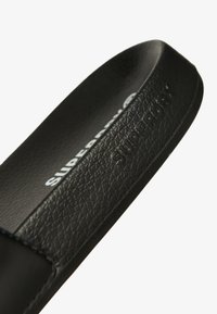 Sandal in pelle sintetica nera con cinturino testurizzato, con il nome del marchio inciso "SUPERDRY" in un colore chiaro a contrasto sulla soletta.