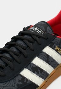 Čierne športové topánky s textúrovaným kvetinovým vzorom, bielymi pruhmi, čiernymi šnúrkami a podrážkou gumovej farby. Interiér má červenú podšívku.