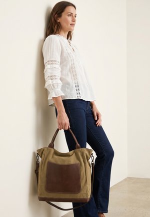Vrouw in witte kanten blouse en donkere spijkerbroek leunt tegen een muur en houdt een grote beige en bruine tas aan de handvatten vast.
