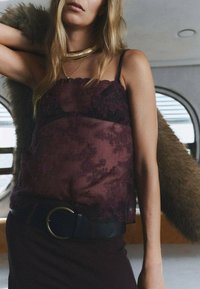 Top burdeos transparente con detalles de encaje, tirantes negros y un collar dorado en capas, combinado con un cinturón negro y un acento de piel marrón.