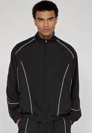Mann trägt schwarze Trainingsjacke mit Reißverschluss und weißen Paspeln, steht mit Händen in den Taschen vor einem schlichten hellen Hintergrund.