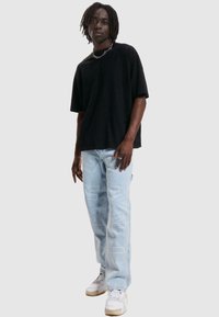 Svart oversized t-skjorte, lyseblå denimjeans med hvit grafisk tekst, og hvite sneakers. Glatt stoff med avslappet passform. Kjedehalskjede detalj.