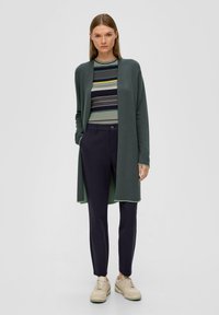 Long gilet à l'avant ouvert de couleur vert foncé, porté par-dessus un haut en tricot rayé dans des nuances de vert, bleu et jaune ; associé à un pantalon bleu marine et des baskets beiges.