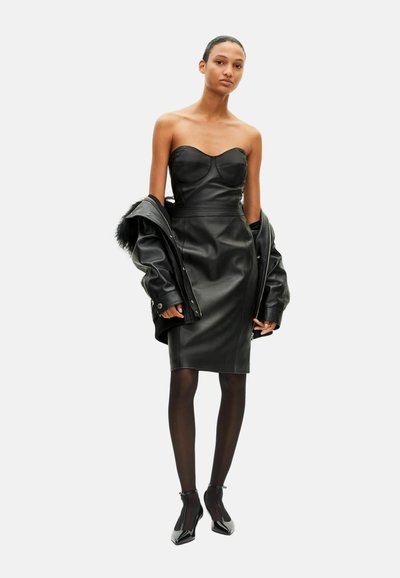 Robe bustier en cuir noir à la silhouette ajustée, associée à une veste noire. Le modèle porte des collants noirs et des escarpins à bout pointu.