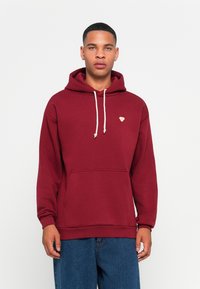 Young man wearing a maroon hoodie with white drawstrings and a small yellow bee patch, paired with blue denim jeans, standing against white background.