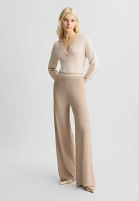 Maglione beige a maglia con scollatura a V profonda e strisce crema sulle maniche, abbinato a pantaloni beige a gamba larga. Scarpe flat dorate a punta.