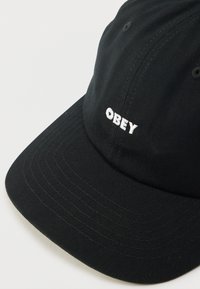 Czarna czapka bejsbolowa wykonana z tkaniny, z zakrzywionym daszkiem, z białym, wyhaftowanym napisem „OBEY” na przodzie w pogrubionej czcionce.