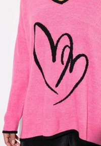 Rosa tröja i mjukt stickat tyg med svarta hjärtformade mönster. Den har en V-ringning och svarta detaljer längs fållen och ärmarna.