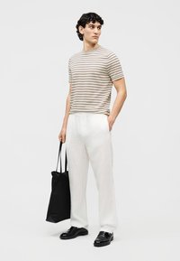 Homme portant une chemise rayée beige et noire, un pantalon blanc, des chaussures noires, tenant un sac fourre-tout noir, se tenant contre un fond clair uni.