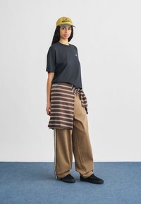 Black t-shirt, brown and white striped wrap, brown wide-leg pants, and a yellow cap; smooth texture, casual design, with black shoes.
