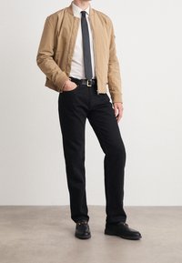 Kaki bomberjacka, vit skjorta, svart slips, svarta jeans, svarta loafers med detaljer i metall. Jackan har ribbade muddar och dragkedja.