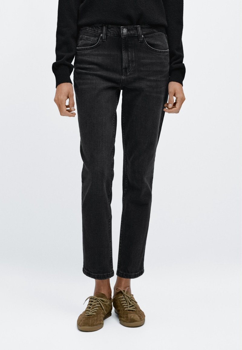 Personne portant un jean noir taille haute et des chaussures en daim marron à lacets, debout devant un fond blanc uni.