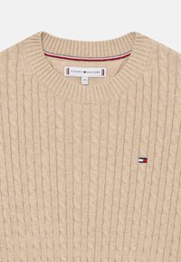 Beige színű, kötött pulóver bordás kerek nyakkal és kicsi piros, fehér és kék logóval a bal mellen. Puha tapintású, jellegzetes mintával.
