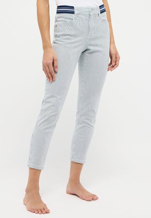 Gros plan d'une personne portant un pantalon à rayures verticales bleu clair et blanc, pieds nus, sur un fond blanc uni.