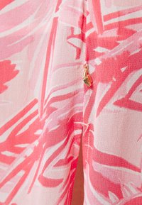 Tissu rose présentant un motif abstrait audacieux dans différentes nuances de rose, avec un petit pendentif banane doré attaché près d'une couture.