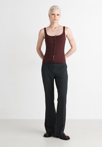 Top senza maniche di color borgogna con chiusura frontale a bottoni, abbinato a pantaloni scuri e strutturati. Tessuto liscio con una forma aderente, modello in posa.