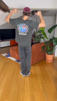 Siva majica s pisanimi grafičnimi motivi na hrbtu, kombinirana s črnimi kavbojkami in belimi supergami. Notranja postavitev s rastlinami.