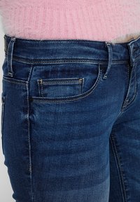 Jeans de mezclilla de color azul oscuro con fit ajustado, con una cintura de altura media, sutiles desgastes y un diseño clásico de cinco bolsillos.