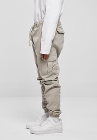 Beige Cargo-Hosen mit elastischen Bündchen, ausgestattet mit Seitentaschen und einem Kordelzug, kombiniert mit weißen Sneakers und einem langärmligen weißen Hemd.