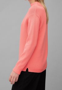 Korallenfarbener gestrickter Pullover mit strukturierter Oberfläche, Rundhalsausschnitt und seitlichen Schlitzen. Lange Ärmel und gerippte Bündchen stehen im Kontrast zum Körper.