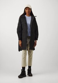 Sieviete, kas valkā bežīgu cepuri, melnu kapuci ilgu jaku, gaiši zilu kreklu, bežīgas īsas bikses, melnas zeķes un melnus zābakus, stāvot pret vienkāršu fona.