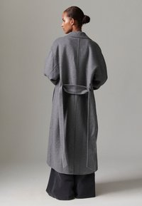 Pilkas per didelis vilnonis paltas su plačiomis rankovėmis, diržu liemenyje ir tiesiu kirpimu. Minkšta tekstūra ir minimalistinis dizainas.