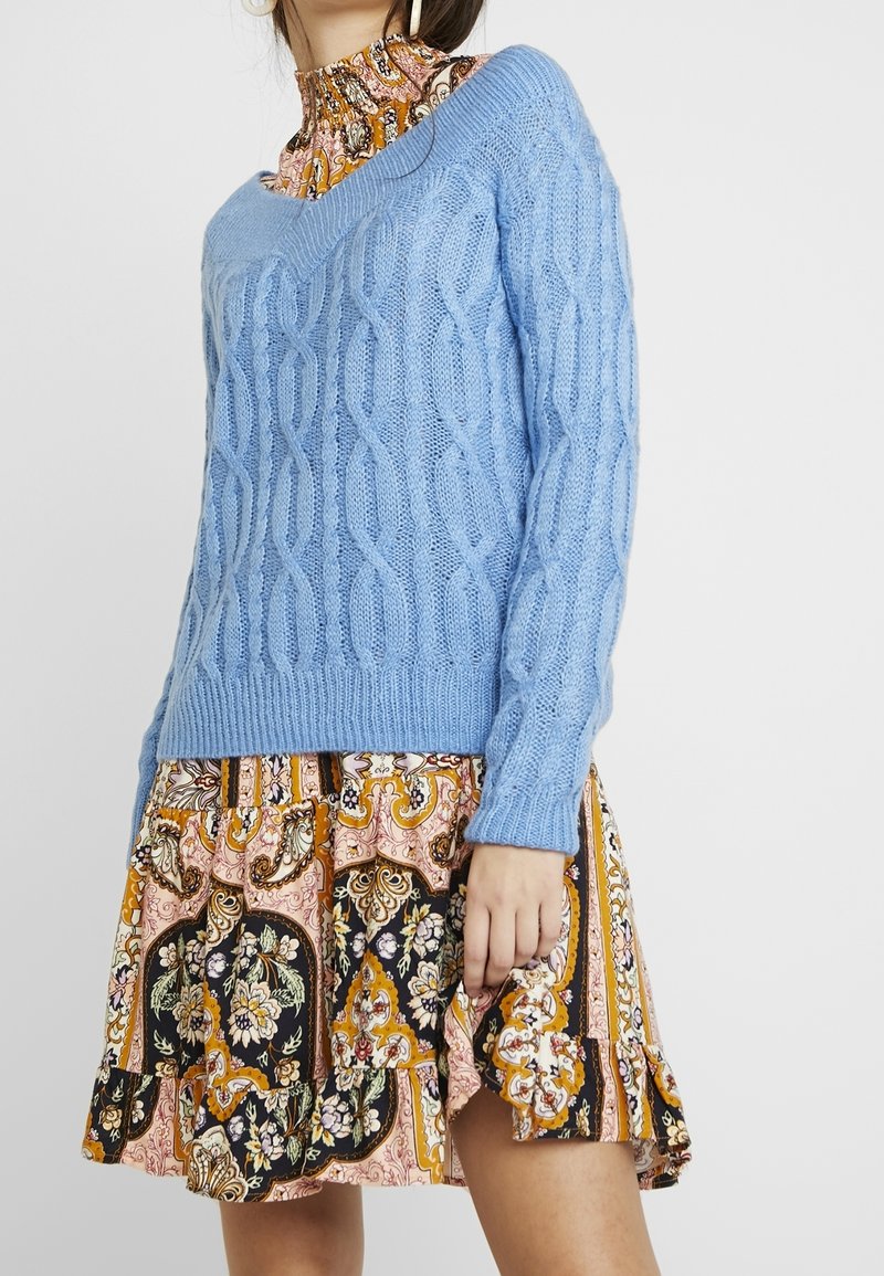 Suéter de punto en azul claro sobre un vestido estampado con diseños florales y de paisley, en tonos de rosa, negro y naranja.