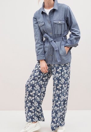 Mujer con chaqueta azul con cinturón, camisa blanca, pantalones de pierna ancha con estampado floral azul y blanco, y zapatillas blancas, sobre un fondo liso.