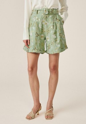 Pantalones cortos de talle alto en verde claro con bordado metálico floral y cinturón a juego, combinados con blusa blanca y sandalias doradas de tacón con tiras.