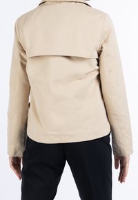 Giacca beige con vestibilità su misura, caratterizzata da un retro piatto e stratificato, maniche lunghe e una superficie liscia e opaca. Abbinata a pantaloni neri.