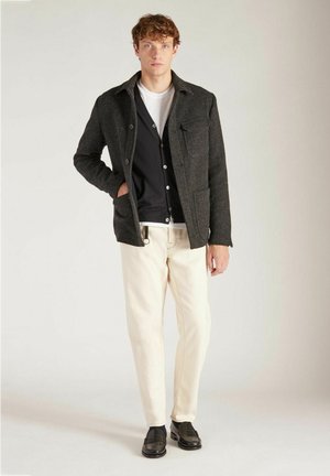 Gray wool jacket with front pockets, black button-up cardigan, white t-shirt, cream pants, and black loafers. Model stands against a light background.