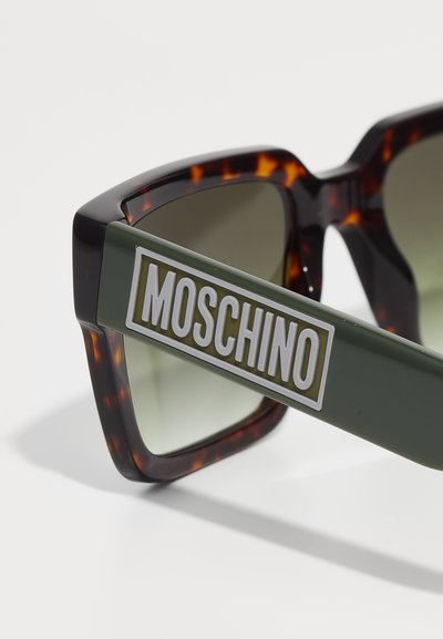 Overdimensjonerte solbriller med et skilpaddemønster på rammen, grønne stenger med en stor hvit "Moschino" logo, og gradient grå linser.