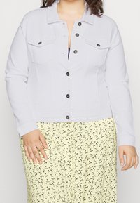 Ljusrgrå jeansjacka med långa ärmar, knäppning, två bröstfickor och svarta knappar. Kombinerad med en gul kjol med blommönster.