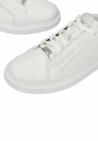 Baskets en cuir blanc avec lacets blancs, panneaux latéraux texturés, plaque métallique sur les lacets et logo sur la languette, présentées sur un fond blanc.