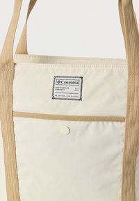 Μπεζ τσάντα ώμου Columbia με ταμπά λαβές, μπροστινή τσέπη ασφαλισμένη με κουμπί κούμπωμα και ραμμένη ετικέτα επωνυμίας με συντεταγμένες στο επάνω μπροστινό πάνελ.