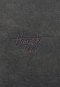 Harmaa tekstiili, jossa on pehmeä rakenne ja tumman violetilla brodeerattu teksti "Rehellisyys Hallitsee" rentouttavassa, käsin kirjoitetussa tyylissä.