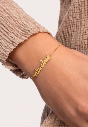 Pulsera de oro con la palabra "abuelo" en una fuente cursiva, acentuada con un pequeño corazón, sobre una muñeca con un fondo de tela texturizada.