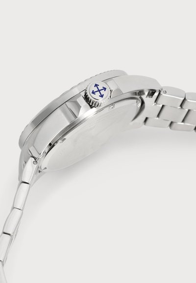 Gros plan de profil d’une montre-bracelet en métal argenté, montrant son boîtier poli, son bracelet à maillons et sa couronne ornée d’un emblème en forme de flèche bleue.