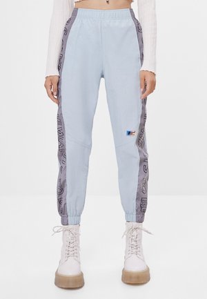 Person wearing light blue jogger pants with gray side panels featuring black text, paired with white lace-up boots and a cropped white top.