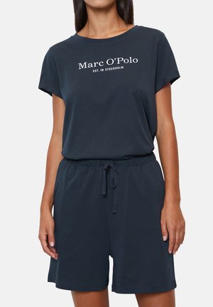 Kvinna i marinblå kortärmad skjorta och matchande shorts med dragsko, skjortan har texten "Marc O'Polo EST. I STOCKHOLM."
