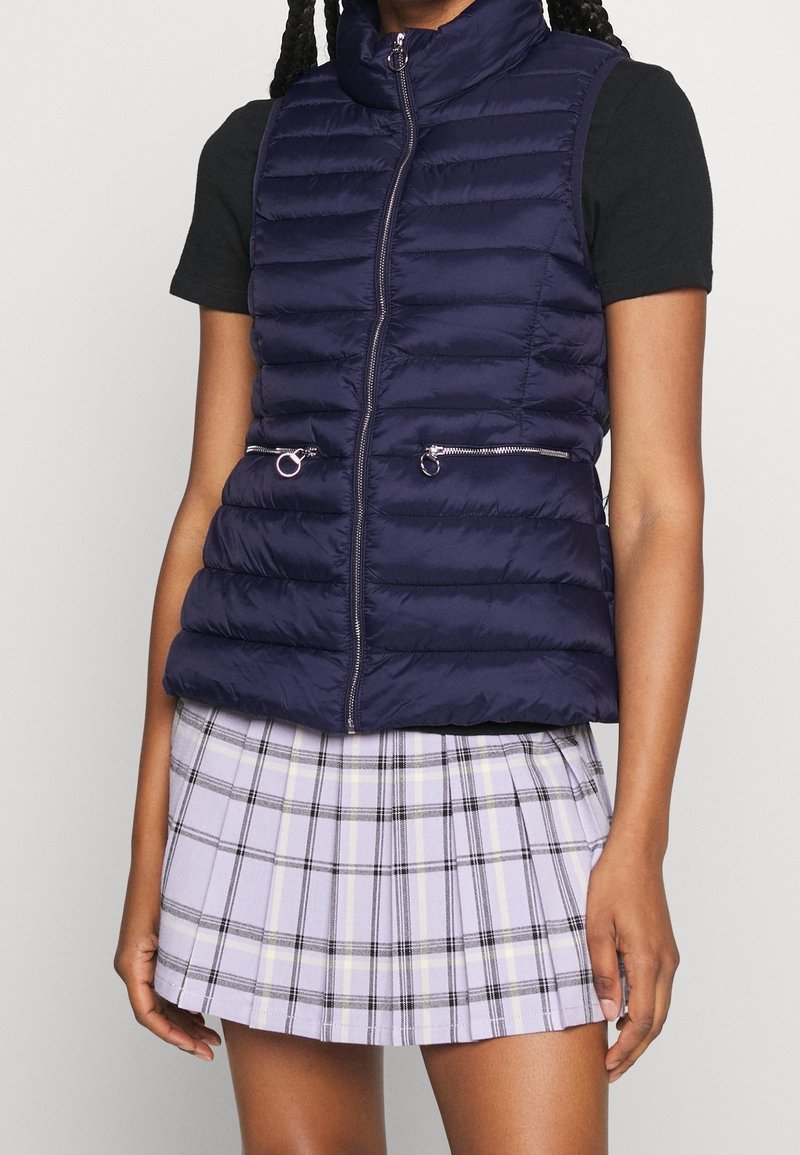 Femme portant un gilet matelassé bleu marine avec des poches zippées sur un t-shirt noir et une jupe plissée à carreaux lavande, debout devant un fond uni.