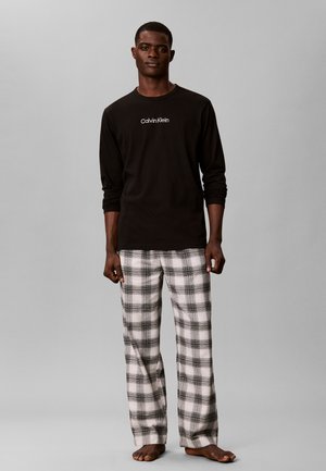 Мъж, стоящ бос, облечен в черна дългорукавна риза Calvin Klein и бели и сиви пижамени панталони на каре на сив фон.